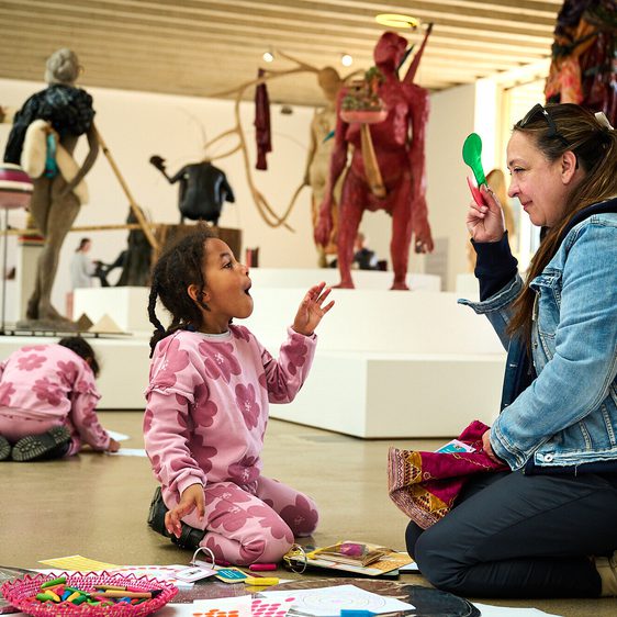 A young girl interacts with a woman seated on the floor, both engaged in a creative activity. In the background, various sculptures are displayed on plinths.