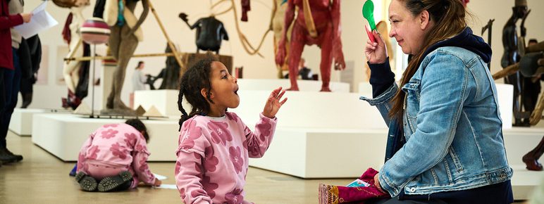 A young girl interacts with a woman seated on the floor, both engaged in a creative activity. In the background, various sculptures are displayed on plinths.