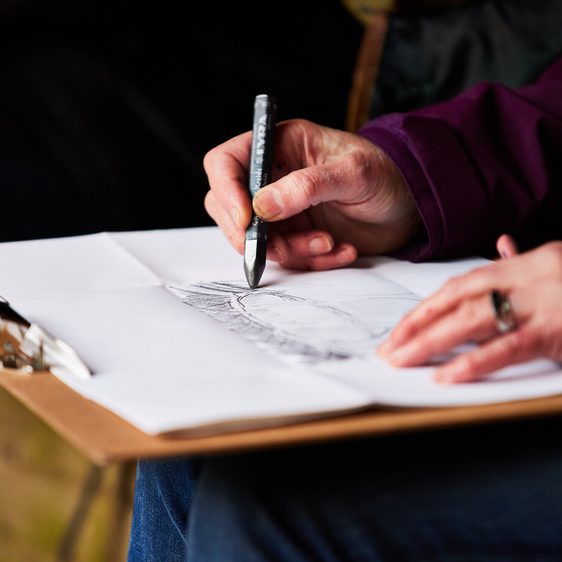 A close-up of a person's hand holding a pen, sketching on a sheet of paper placed on a clipboard.