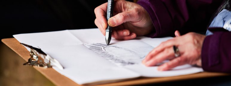 A close-up of a person's hand holding a pen, sketching on a sheet of paper placed on a clipboard.