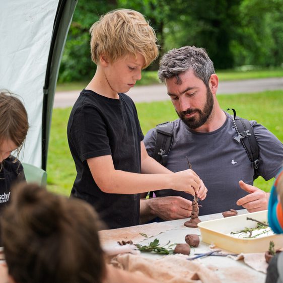 A group of children and an adult are engaged in a clay crafting activity outdoors. A boy is focused on shaping a piece of clay, while a man observes and assists.