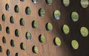 A close-up of a rusted metal panel featuring evenly spaced circular holes. The reflections in the holes show greenery, creating a contrast between the textured surface and the vibrant natural surroundings.
