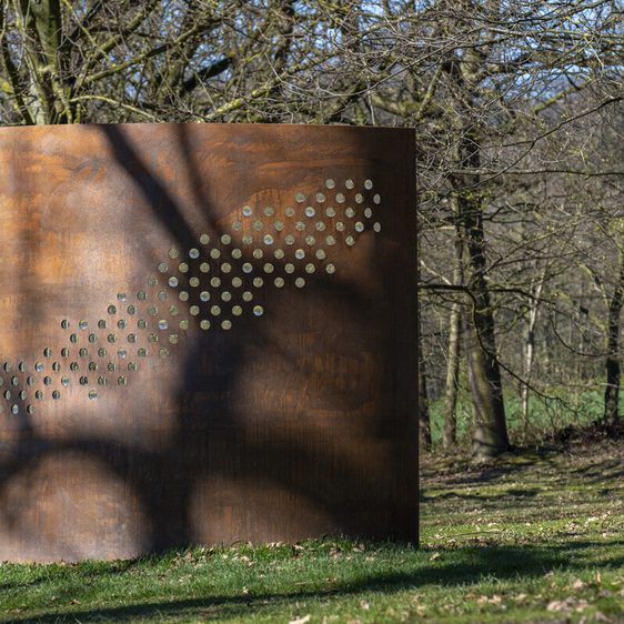 A large, rusted metal sculpture stands in a wooded area, featuring a series of circular cut-outs that create a pattern. Sunlight filters through the trees, casting shadows on the sculpture and the grassy ground around it.