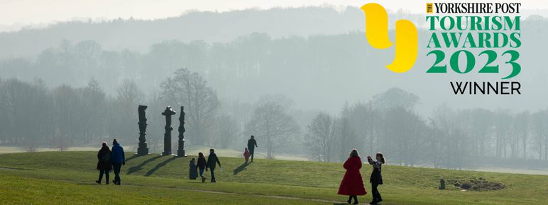 People walking across a hillside, with three tall thin silhouetted sculptures and trees in the background. The Yorkshire Post Tourism Award 2023 Winner logo is in the top right corner.