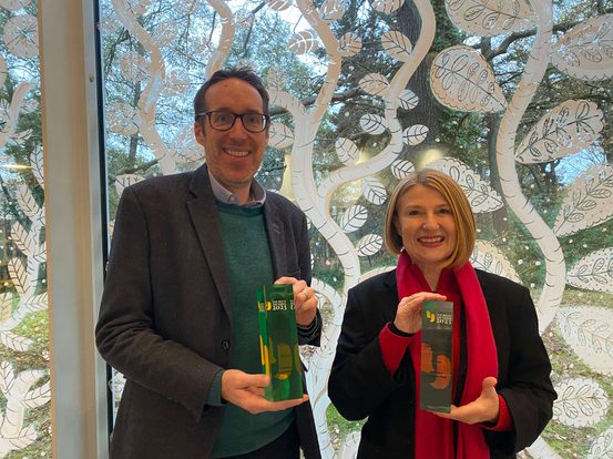 A smiling man and woman holding rectangular glass awards in front of a window decorated with white painted leaves