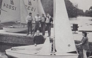 Four boys wait on the dock by the lake to get in to the sail boat held by two adults.