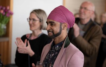 A man wearing a turban smiling with a champagne glass in his hand and a woman clapping behind him.