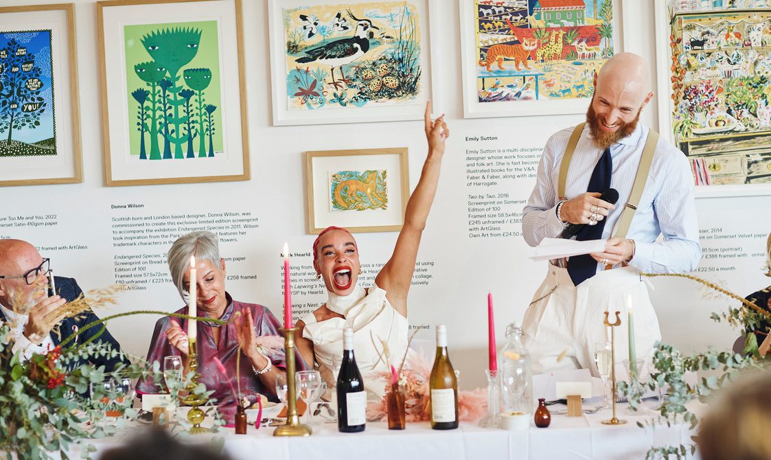 A wedding party during speeches. They are seated at a table decorated with candles and flowers, with framed prints behind them.