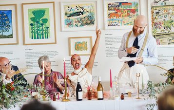 A wedding party during speeches. They are seated at a table decorated with candles and flowers, with framed prints behind them.
