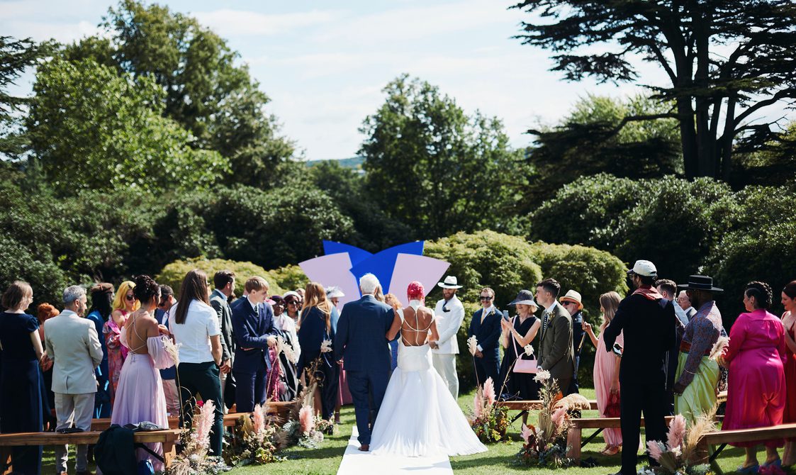 A bride and escort, walking down an outdoor aisle surrounded by their guests.
