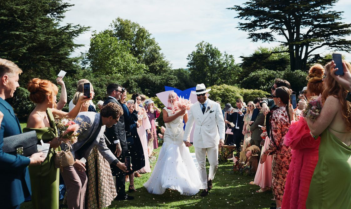 A bride and groom, wearing a long white dress and white suit, walking down an outdoor aisle surrounded by their guests.