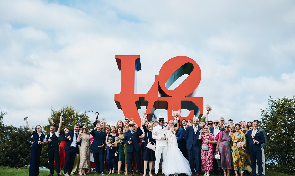 A wedding party, including the bride and groom and guests, standing in front of a large red LOVE sculpture outdoors.