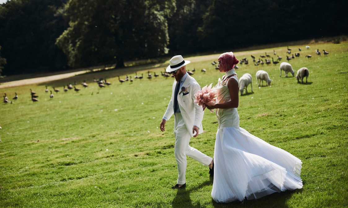 A bride and groom, wearing a long white dress and white suit, walking outdoors through a field of sheep and geese