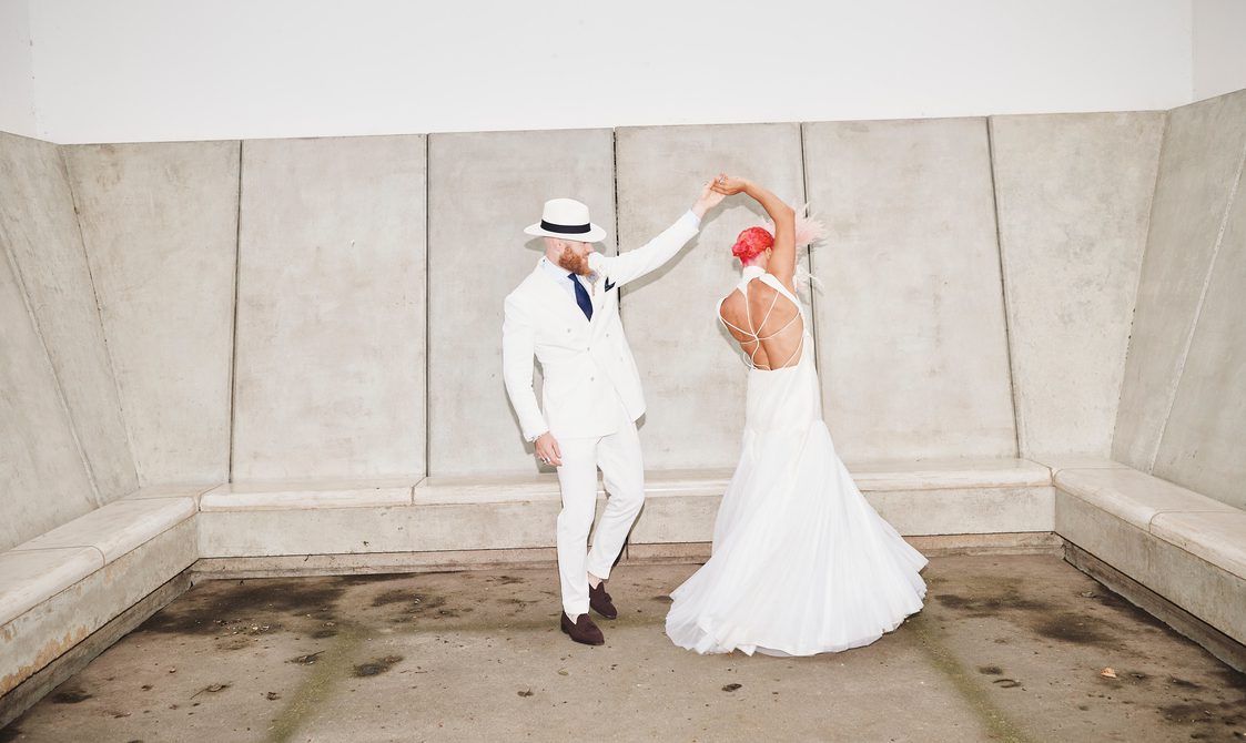 A bride and groom, wearing a long white dress and white suit, dancing inside a small room with concrete seating.