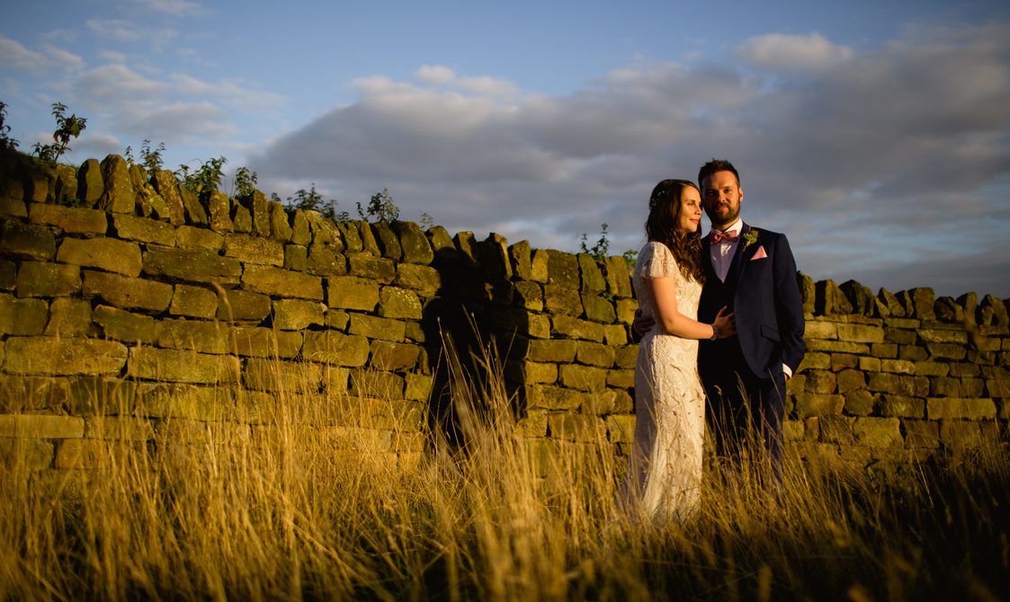 A bride and groom standing in front of a dry stone wall at sunset