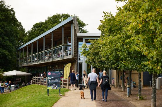 A couple with a dog on a lead, walking towards the YSP Visitor Centre, a modern angular building, with a balcony.