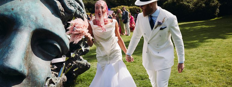 A bride and groom, wearing a white structured wedding dress and white suit, holding hands while walking past a giant bronze head sculpture.