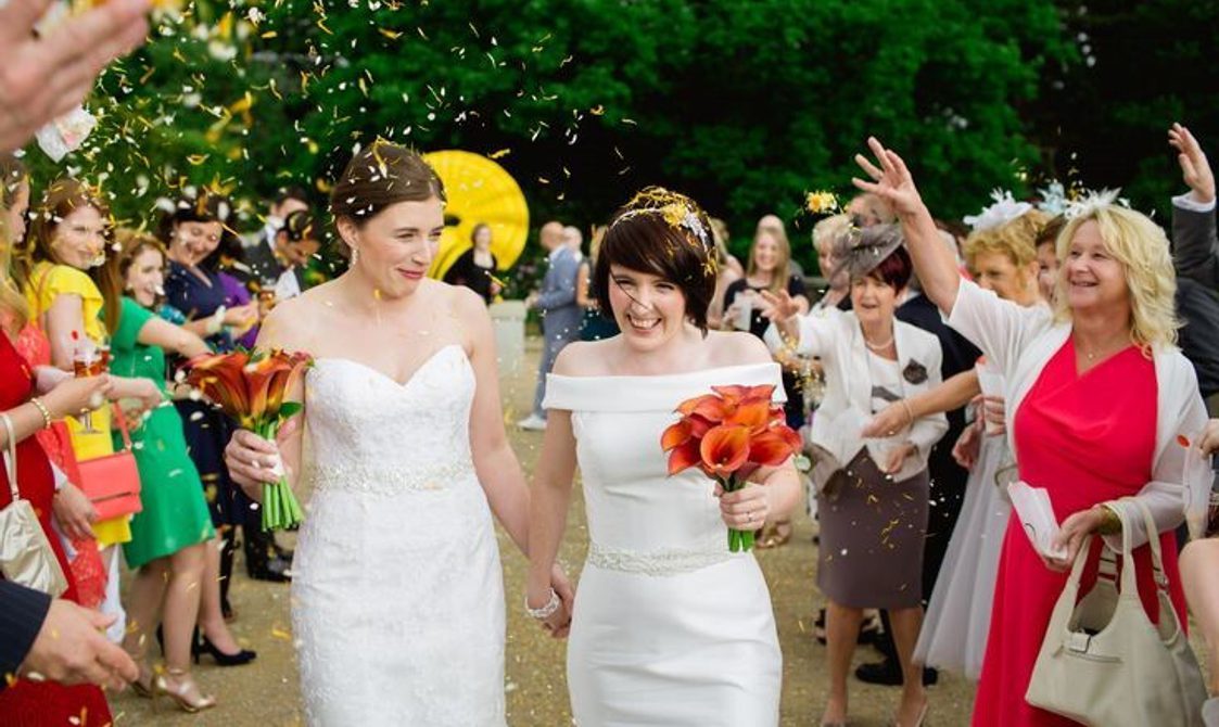 A pair of brides in white wedding dresses, walk between two rows of friends and family throwing petal confetti.