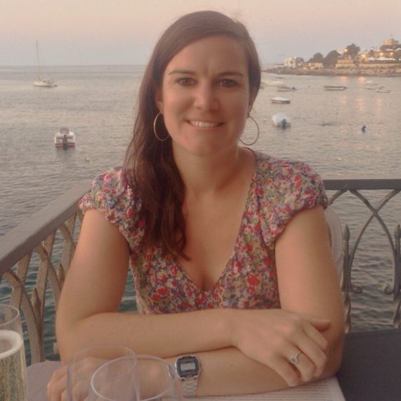 A woman with long brown hair sitting at a table with the sea behind her