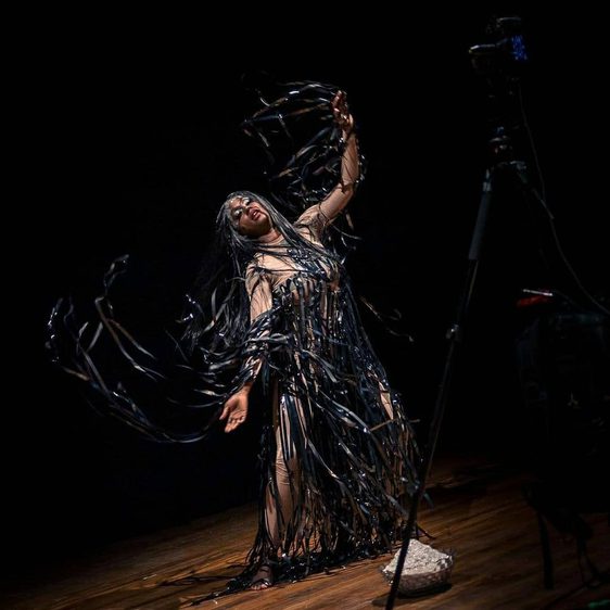 A black woman with her arms in the air, wearing a costume of long black strings