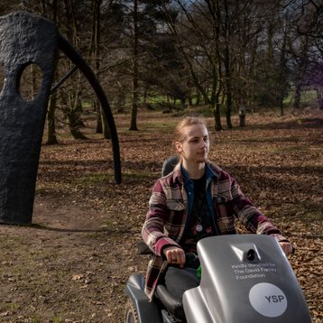 Visitor passing a sculpture in the parkland on a mobility scooter at YSP