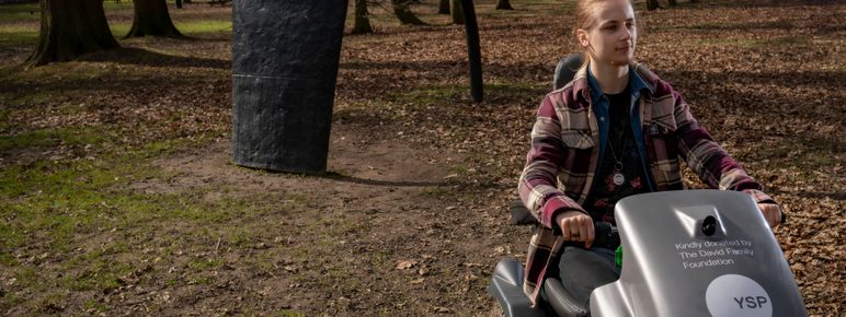 Visitor passing a sculpture in the parkland on a mobility scooter at YSP