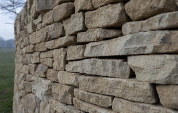 A close-up view of a dry stone wall made of irregularly shaped, weathered stones. The wall features a mix of light and dark hues, showcasing the texture and natural patterns of the stones against a blurred green landscape in the background.