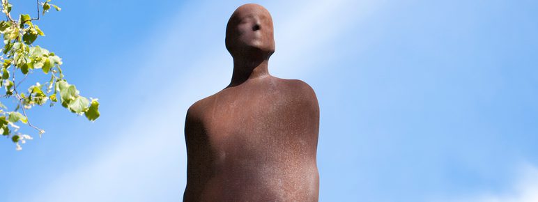 A sculpture of a male figure, displayed on a dead tree trunk with blue sky and clouds behind it.