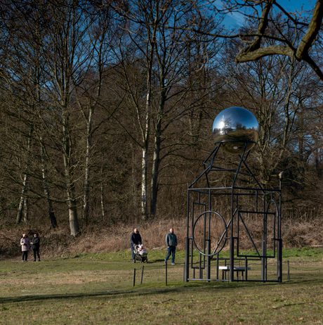 People with a pushchair walking past a metal frame sculpture with a silver ball at the top.