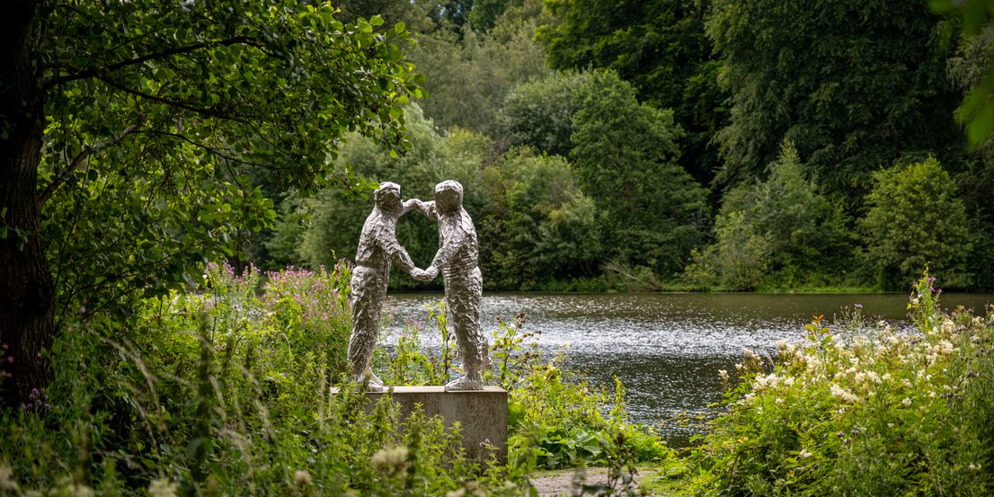 A silver sculpture of two figures dancing. A lake, trees and foliage surround it.