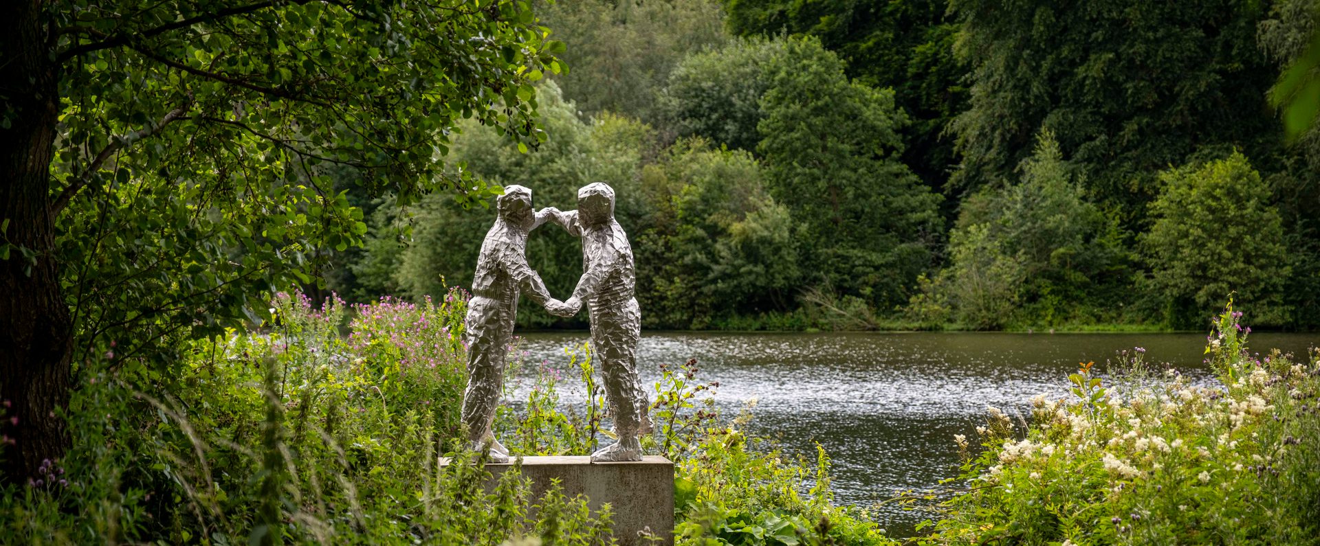 A silver sculpture of two figures dancing. A lake, trees and foliage surround it.