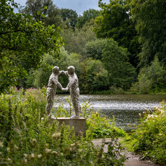A silver sculpture of two figures dancing. A lake, trees and foliage surround it.