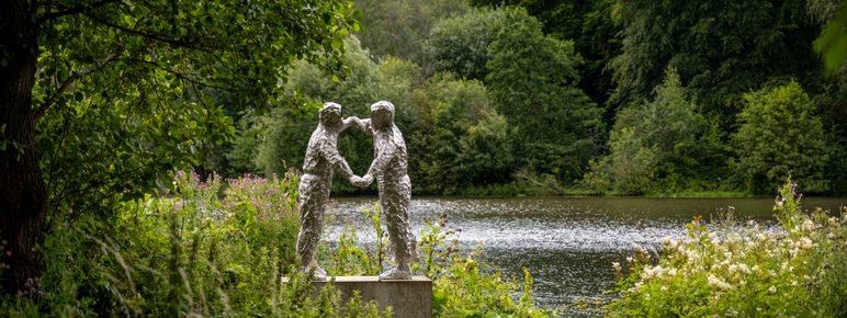 A silver sculpture of two figures dancing. A lake, trees and foliage surround it.