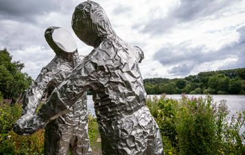 Two silver figures in astronaut suits with flat expressionless faces, in an embrace. A lake is behind them.