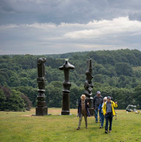People in raincoats walking past three tall thin bronze sculptures looking out over the landscape