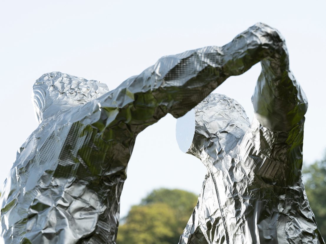 Two silver figures in astronaut suits with flat expressionless faces, in an embrace.