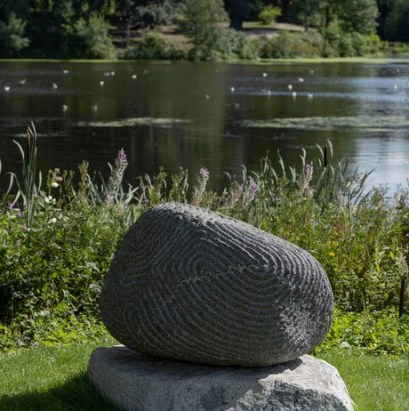 A granite sculpture with intricate carved lines in front of the Lower Lake in sunshine
