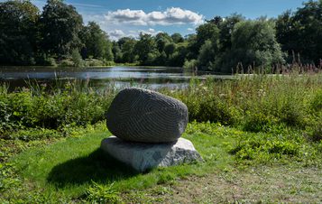 A granite sculpture with intricate carved lines in front of the Lower Lake in sunshine