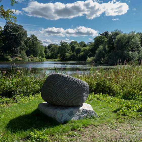A granite sculpture with intricate carved lines in front of the Lower Lake in sunshine