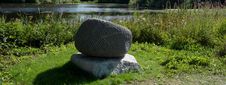 A granite sculpture with intricate carved lines in front of the Lower Lake in sunshine