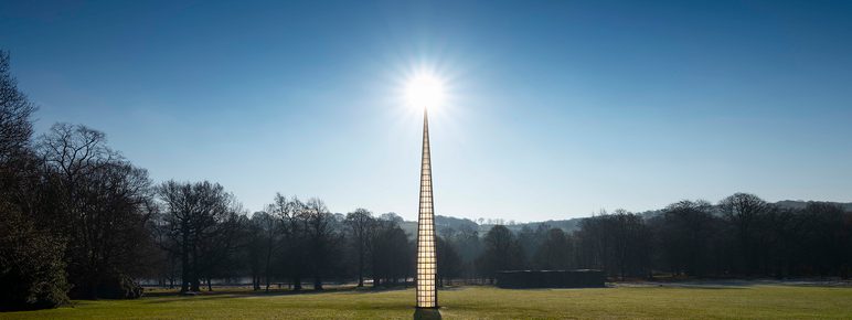 A tall pointed tower, with glass panels. The sun is rising behind it.