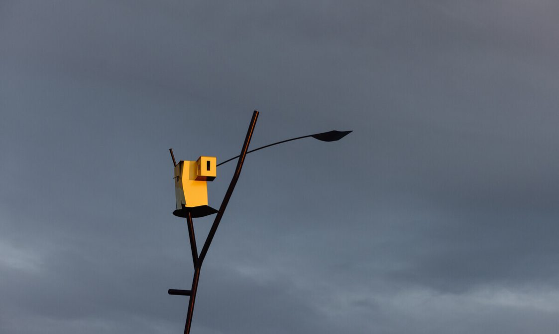 A tall, slender sculpture stands on a grassy hill, featuring a unique birdhouse design at its top. The sky is overcast with soft grey clouds.