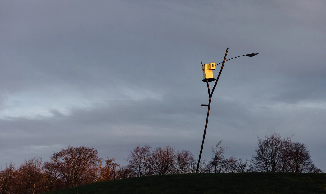 A tall, slender sculpture stands on a grassy hill, featuring a unique birdhouse design at its top. The sky is overcast with soft grey clouds.