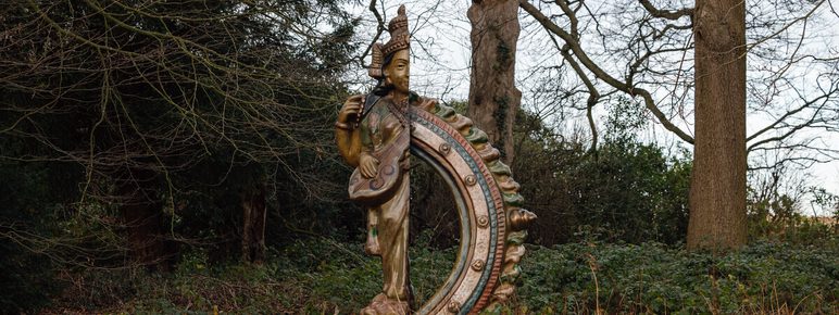 A bronze sculpture of a human figure in traditional South Asian dress, with half replaced by a decorative curved section. It is displayed outdoors in woodland.