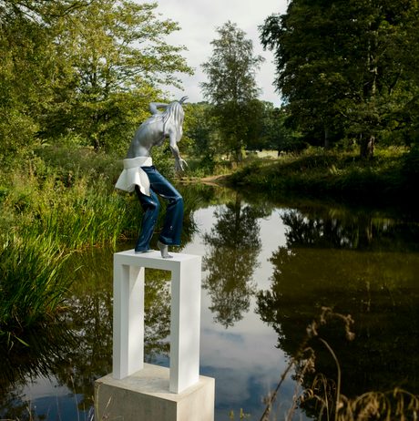 A female figure wearing jeans standing on a white plinth at the edge of a lake, surrounded by foliage
