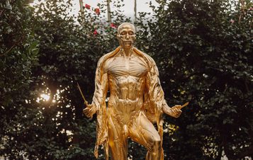 A gilded gold sculpture of a human figure with the skin removed revealing the musculature, displayed inside a glass house with large bushes. The figure is holding a knife and scissors.