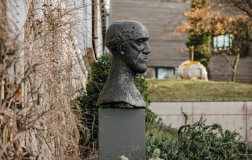 A bronze bust of a human head displayed on a tall grey plinth outdoors with climbing plants behind it.