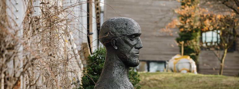 A bronze bust of a human head displayed on a tall grey plinth outdoors with climbing plants behind it.