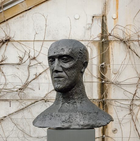 A bronze bust of a human head displayed on a tall grey plinth outdoors with climbing plants behind it.