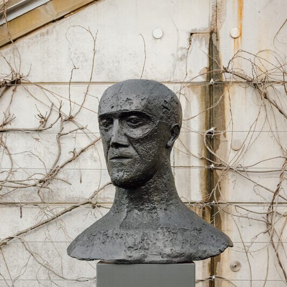A bronze bust of a human head displayed on a tall grey plinth outdoors with climbing plants behind it.